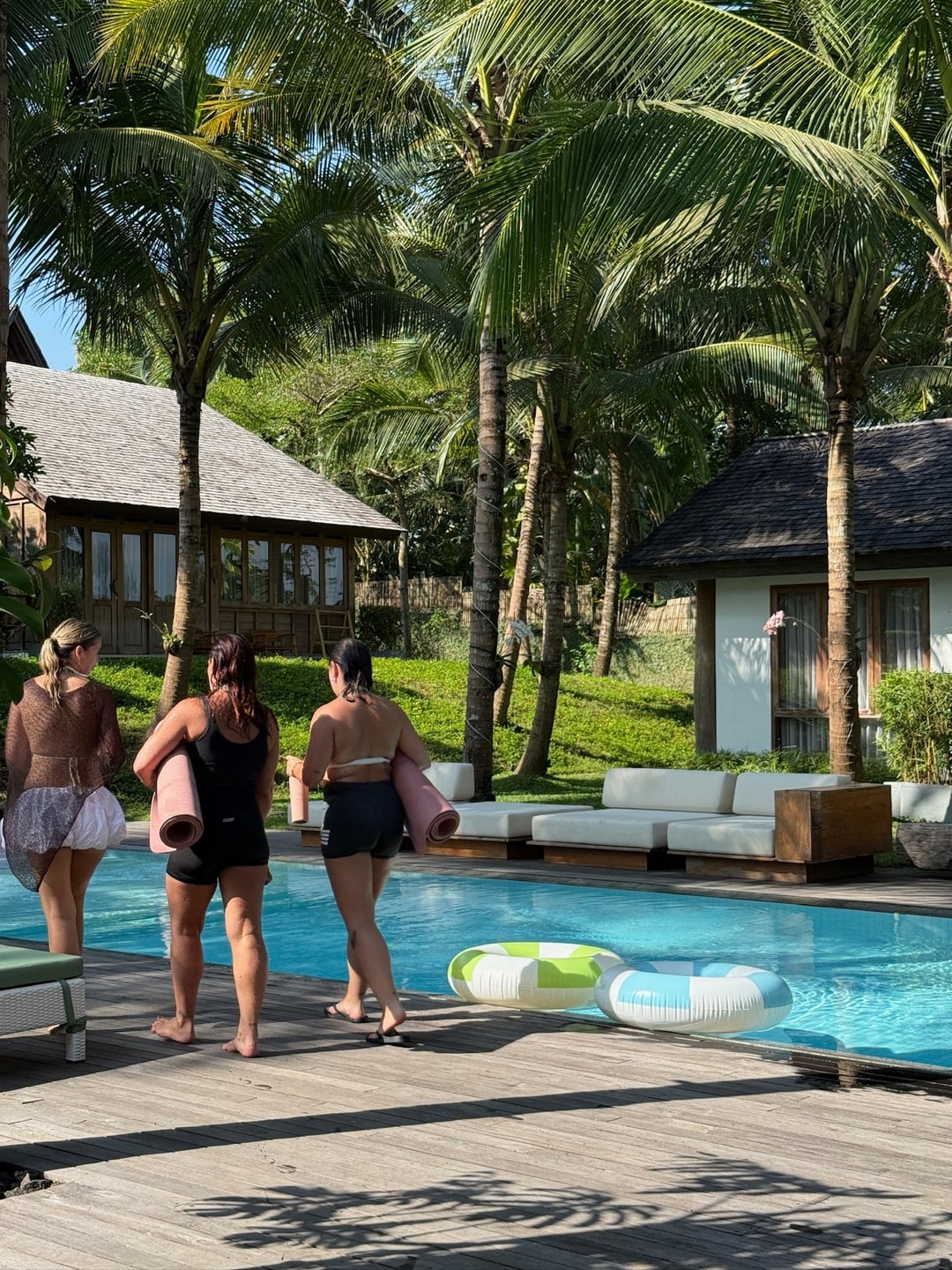 Three retreat participants carrying rolled exercise mats beside a pool surrounded by palms
